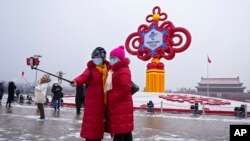 Gratë që mbajnë maska për fytyrën të printuara me një slogan për Lojërat Olimpike Dimërore të Pekinit bëjnë një selfie me një dekorim të lojërave në sfond, në sheshin Tiananmen në Pekin, 20 janar 2022 (AP Photo/Andy Wong)