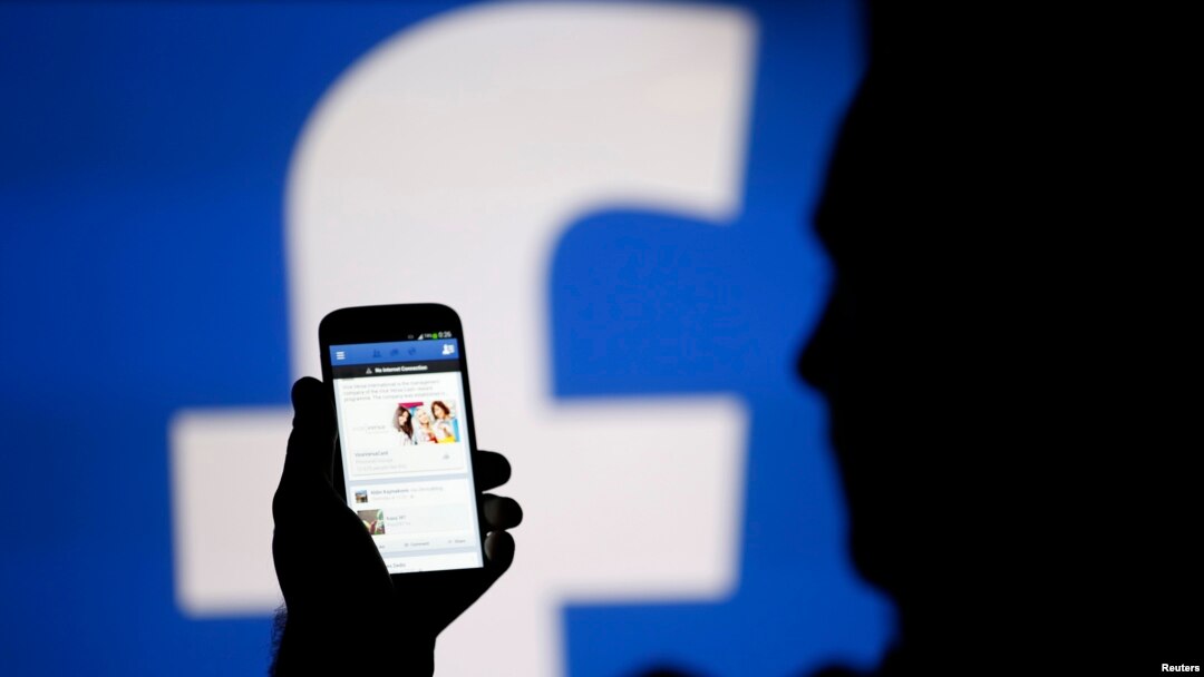 A man is silhouetted against a video screen with a Facebook logo holding a smartphone in the central Bosnian town of Zenica, August 14, 2013.