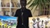 Santo Makoi poses in front of two of his recent paintings of village life in South Sudan. He says that he likes to show people what life is like back home.