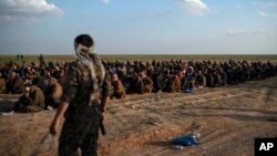 FILE - U.S.-backed Syrian Democratic Forces fighters stand guard next to men waiting to be screened after being evacuated out of the last territory held by Islamic State group militants, near Baghuz, eastern Syria, Feb. 22, 2019.