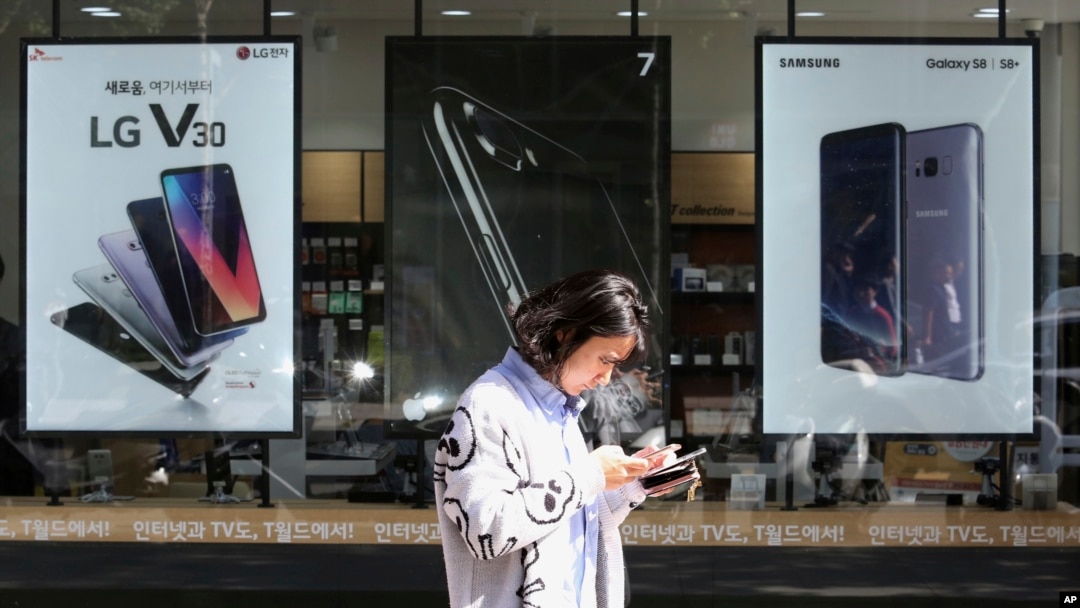FILE - A woman checks her phone as she walks by posters adverting smartphones at a mobile phone store in Seoul, South Korea, Oct. 17, 2017.